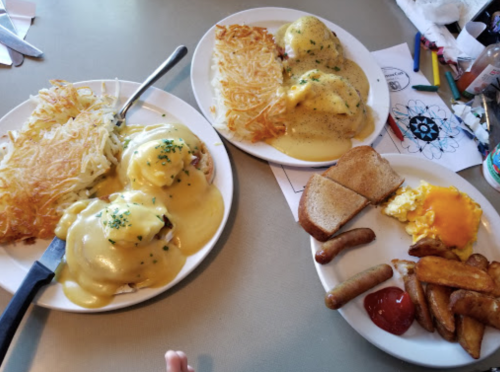 Two plates of breakfast food featuring eggs Benedict, hash browns, toast, sausage, and a side of cheese.