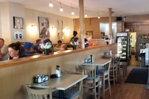 A cozy café interior with wooden tables, patrons dining, and a counter displaying food and drinks. Warm lighting enhances the atmosphere.