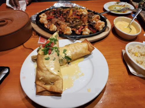 A plate with a crispy rolled dish topped with cheese and herbs, alongside a sizzling platter of shrimp and vegetables.