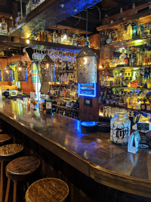 A vibrant bar with wooden stools, colorful bottles on shelves, and decorative lighting creating a lively atmosphere.