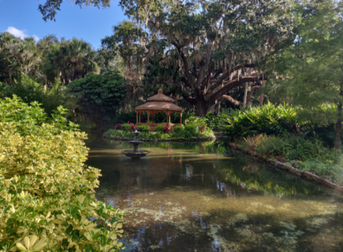 A serene garden with a gazebo surrounded by lush greenery and a calm pond reflecting the trees.