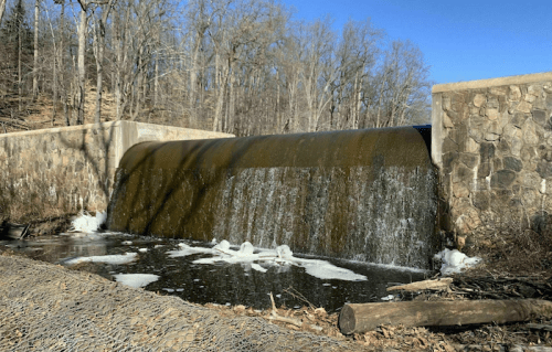 A stone dam with water cascading over it, surrounded by trees and ice on the water's surface. Clear blue sky above.