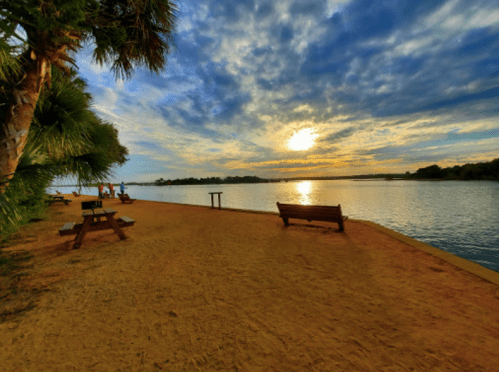 A serene riverside scene at sunset, featuring benches, palm trees, and a colorful sky reflecting on the water.