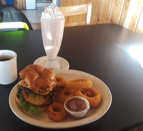 A plate with a burger, onion rings, and a small cup of ketchup, alongside a pink milkshake and a coffee cup.