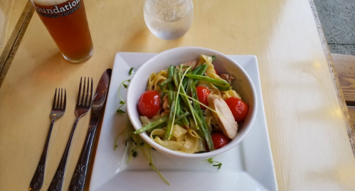 A bowl of pasta with chicken, cherry tomatoes, and green beans, alongside a glass of water and a beer on a table.