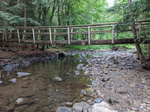 A wooden bridge spans a rocky stream surrounded by lush green trees in a serene forest setting.