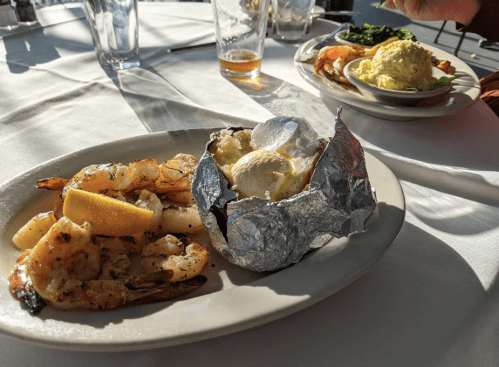 A plate with grilled shrimp and a baked potato wrapped in foil, with a lemon wedge. Another plate with shrimp and mashed potatoes in the background.