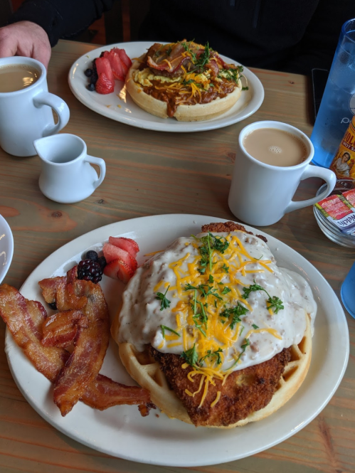 A plate of chicken and waffles topped with cheese and gravy, served with bacon and fresh fruit, alongside coffee.