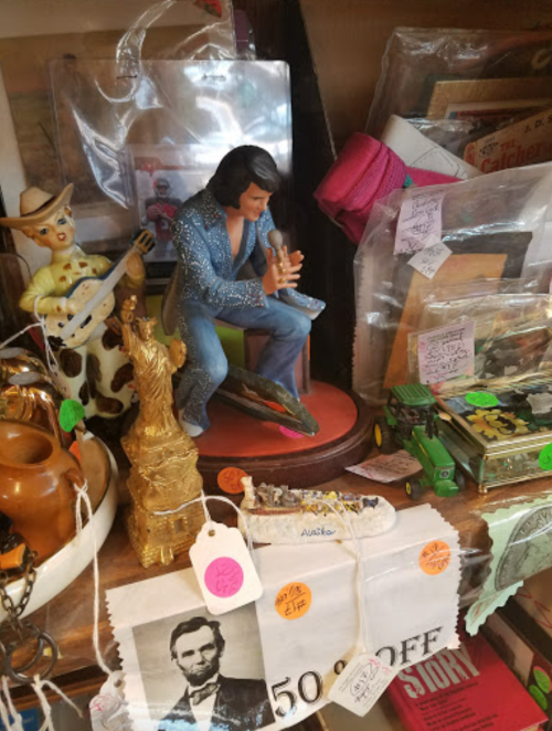 A cluttered display of collectibles, featuring an Elvis figurine, a cowboy, and various vintage items with price tags.