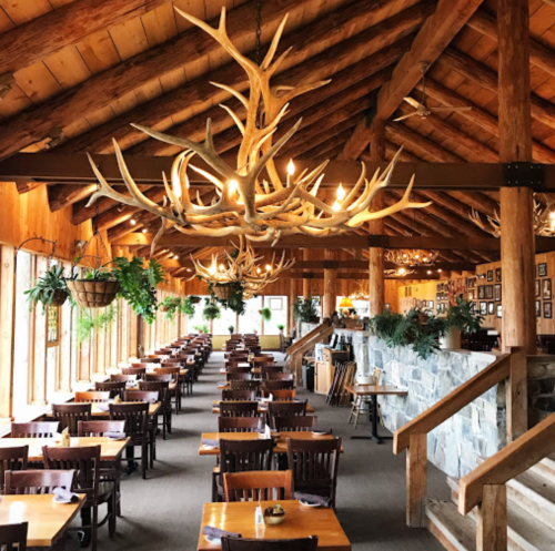 Rustic restaurant interior with wooden beams, antler chandeliers, and neatly arranged tables. Greenery adds a cozy touch.