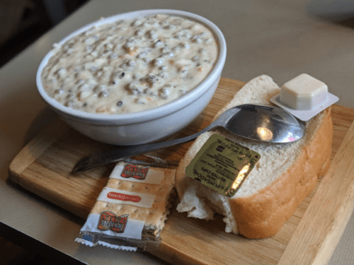 A bowl of creamy soup with meat and vegetables, served with a slice of bread and butter on a wooden board.