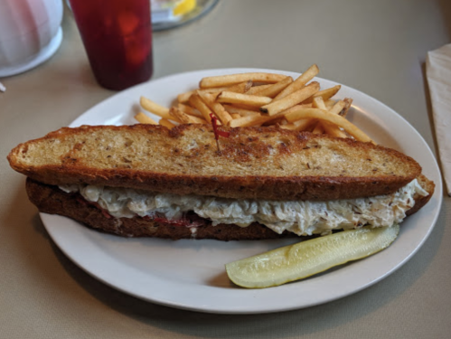 A large sandwich with a crispy crust, filled with coleslaw, served with fries and a pickle on the side.
