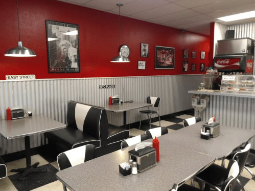 A retro diner with red walls, black and white seating, and checkered floors, featuring tables and a counter display.