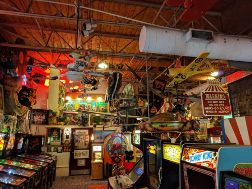 A vibrant arcade filled with vintage pinball machines, colorful decor, and whimsical hanging objects.