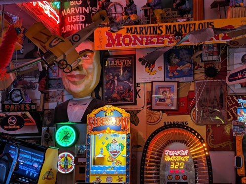 A vibrant, eclectic wall filled with vintage signs, arcade games, and a large clown head in a lively setting.