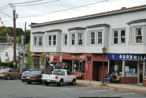 A small town scene featuring diners, a Goodwill store, and parked cars near a railroad crossing.