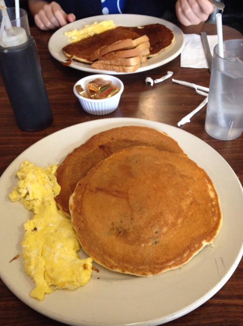 A plate with two large pancakes, scrambled eggs, and toast, with syrup and drinks in the background.