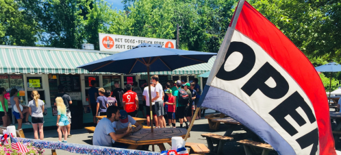 A busy outdoor eatery with a large "OPEN" flag, people enjoying food, and a sunny day in the background.