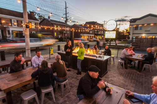 Outdoor dining scene at dusk with people socializing around tables and a fire feature, string lights overhead.