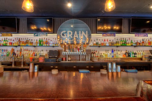A bar with a wooden counter, shelves filled with various bottles, and a sign reading "GRAIN" in the center.