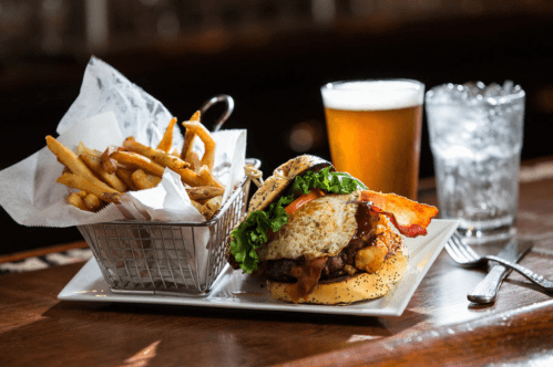 A juicy burger topped with bacon, lettuce, and a fried egg, served with fries and drinks on a wooden table.