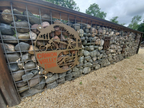 Sign for the Cannon Valley Trail mounted on a stone wall, surrounded by gravel and greenery.
