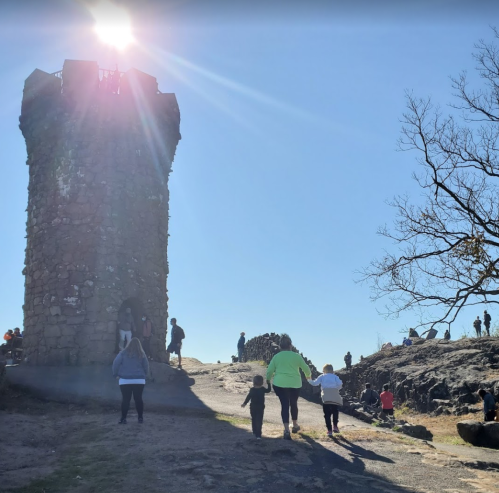 A stone tower on a rocky hill with people walking nearby and others gathered at the top, under a bright sun.