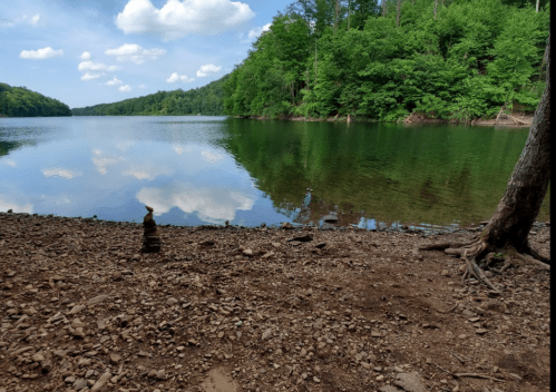 A serene lake surrounded by lush greenery, with a stack of stones on the shore and reflections in the water.