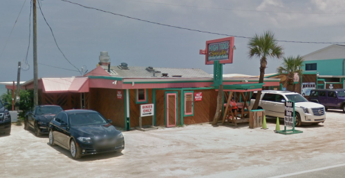 A beachside restaurant with a wooden exterior, parking for bikes, and palm trees nearby under a clear sky.
