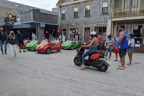 A woman on a scooter rides past a group of people near colorful go-karts in a lively street setting.