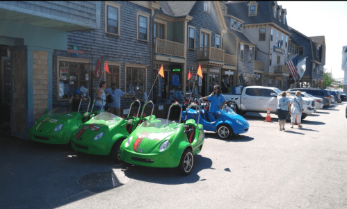 Colorful go-karts parked along a street with shops, people walking, and flags in a sunny outdoor setting.