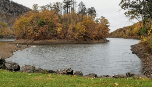 A serene lake surrounded by autumn trees, with people fishing along the shore and rocky terrain in the foreground.