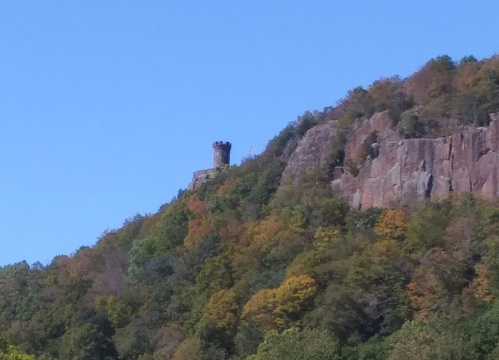 A stone tower atop a rocky hill, surrounded by trees displaying autumn colors under a clear blue sky.