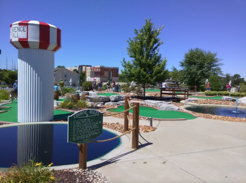 A mini-golf course with colorful structures, water features, and a sign displaying course rules under a clear blue sky.