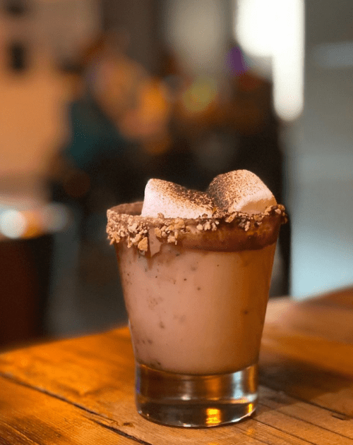 A creamy drink in a glass, topped with marshmallows and chocolate, sitting on a wooden table.