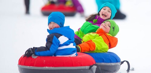 Three children joyfully tubing down a snowy hill, wearing colorful winter gear and laughing together.