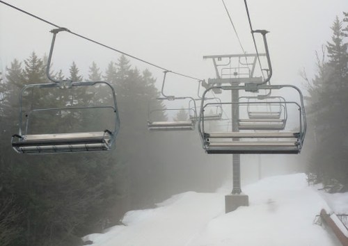 Empty ski lift chairs suspended above a snowy path, surrounded by fog and trees.
