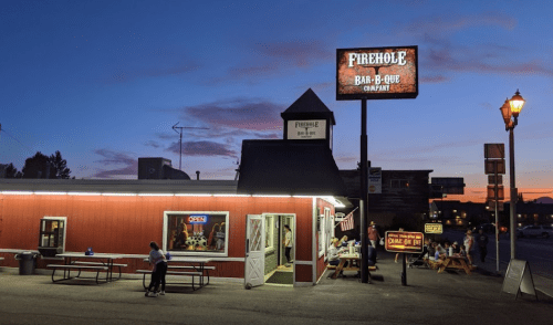 A barbecue restaurant at dusk, with outdoor seating and a glowing "Open" sign. The sky is colorful with sunset hues.