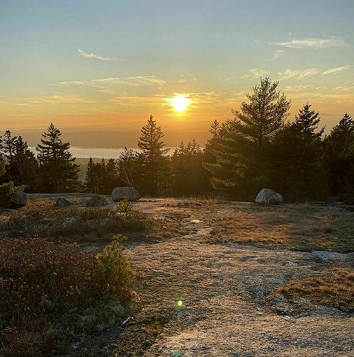 Sunset over a rocky landscape, with trees silhouetted against a colorful sky and distant water visible.
