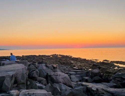 A person sits on rocky shore, watching a serene sunset over calm waters, with vibrant orange and purple hues in the sky.