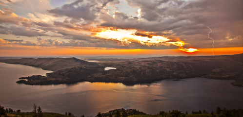 A dramatic sunset over a lake, with clouds and a lightning strike illuminating the sky.