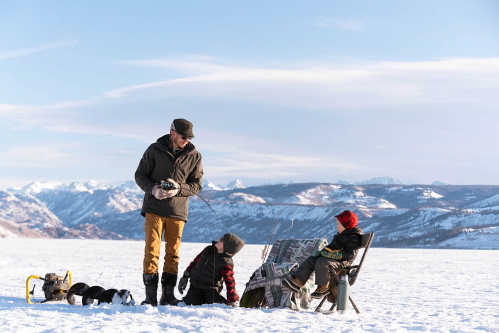 A man stands on a snowy landscape with two children, one sitting in a chair, while ice fishing gear is nearby.