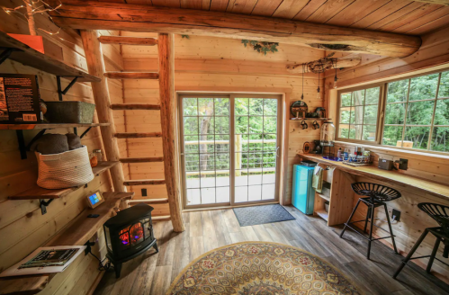 Cozy wooden interior of a cabin with a small kitchen area, a fireplace, and large windows overlooking greenery.