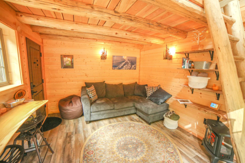 Cozy wooden interior of a small cabin featuring a sectional sofa, decorative shelves, and warm lighting.