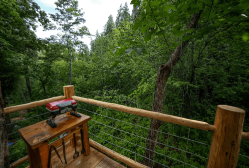A wooden balcony overlooking a lush green forest, with a small grill and tools on a table.