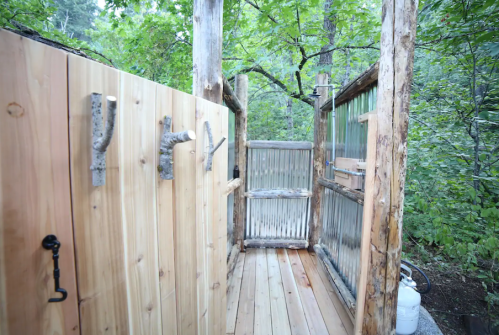 Outdoor shower area surrounded by wooden walls and greenery, featuring rustic hooks and a wooden floor.