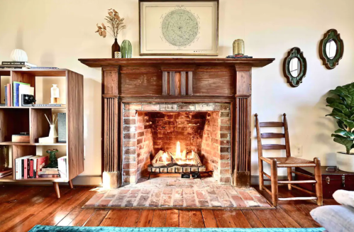 Cozy living room with a brick fireplace, wooden mantel, bookshelves, and decorative mirrors. Warm and inviting atmosphere.