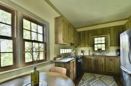 Cozy kitchen with wooden cabinets, large windows, and a round dining table, featuring a patterned rug and modern appliances.