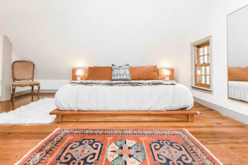 Cozy bedroom with a wooden bed, decorative pillows, a rug, and soft lighting, featuring a minimalist design.