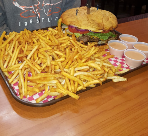 A large burger with toppings sits on a tray surrounded by a mountain of fries and four dipping sauces.